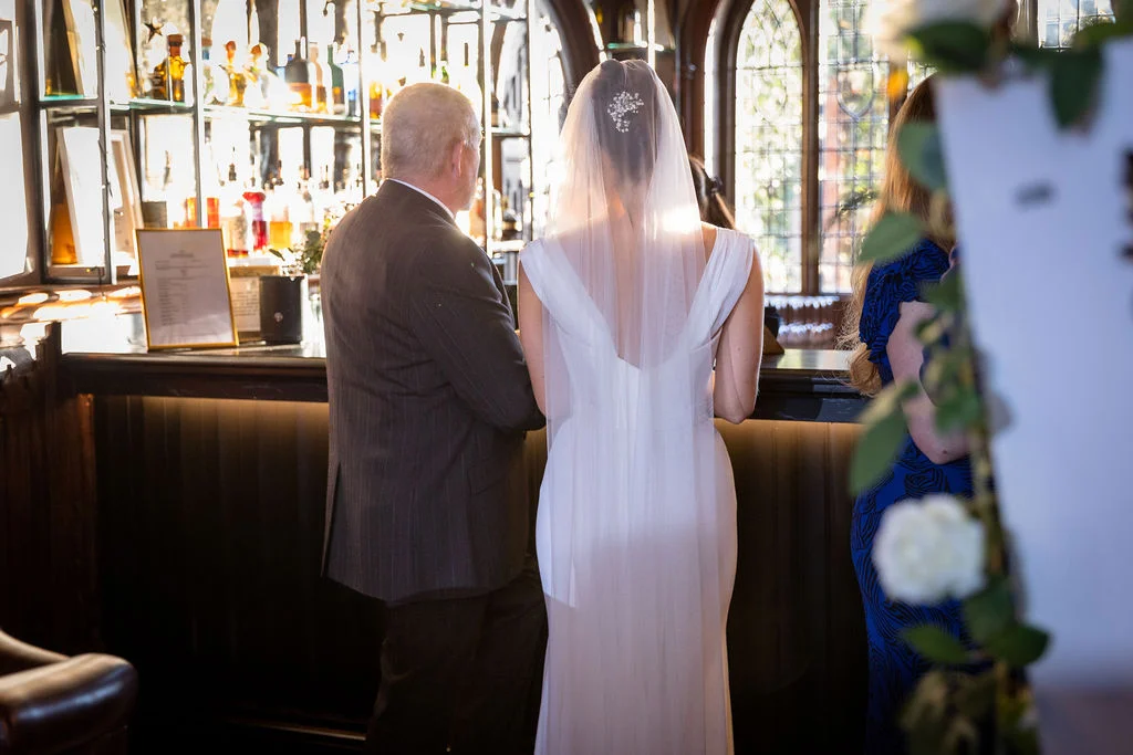 A bride in a white dress with a veil stands with her father at a bar inside a pub wedding venue.