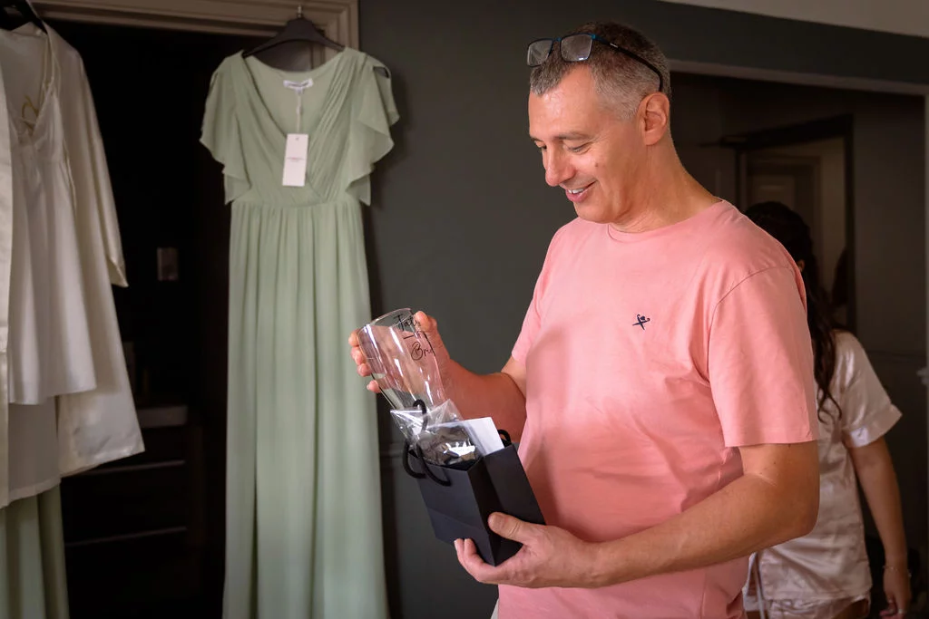 A smiling father of the bride examines a gift while getting ready indoors near a hanging sage green dress.