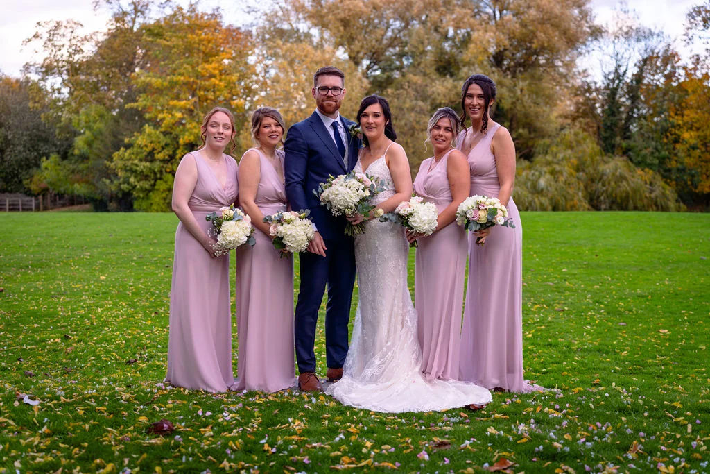 A newlywed couple poses outdoors on a lush green lawn surrounded by bridesmaids in floor-length pink dresses holding bouquets, with autumn trees in the background.