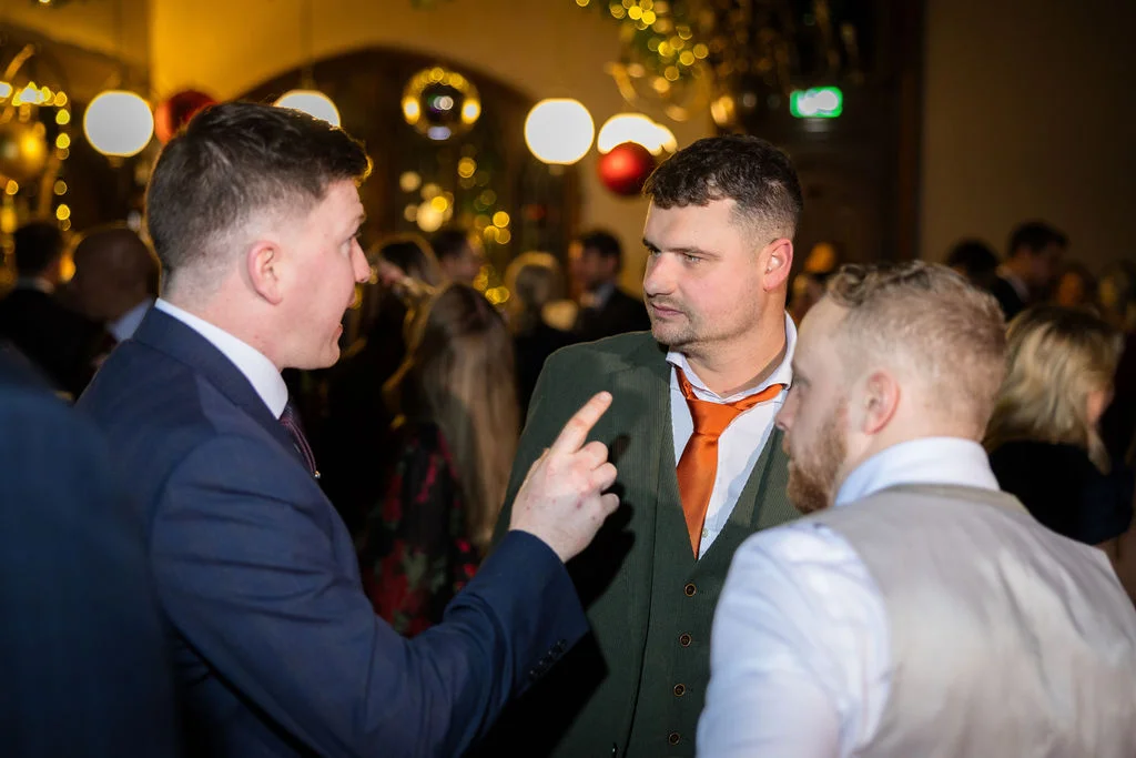 Several wedding guests dressed in sharp suits chat animatedly during an evening reception illuminated by warm, glowing overhead lights.