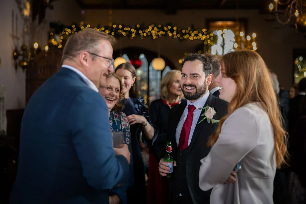 A smiling groom holding a beer bottle chats and laughs with wedding guests during a lively indoor reception under a canopy of fairy lights.