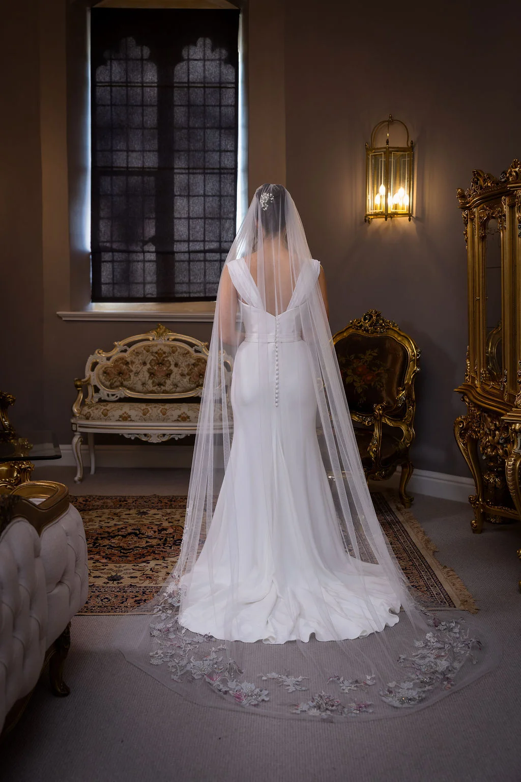 A bride stands facing a large window in a stately manor house, wearing an elegant crepe wedding dress with a long embroidered veil and button-back detail.