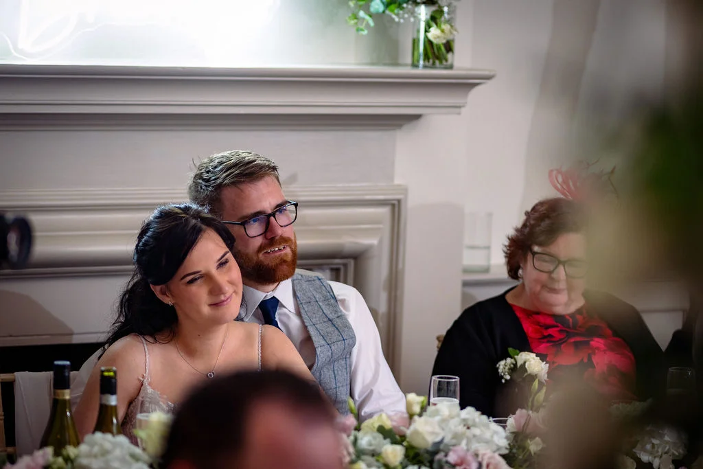 A bride and groom smile while sitting at the top table during wedding speeches, with a guest in a fascinator seated nearby.