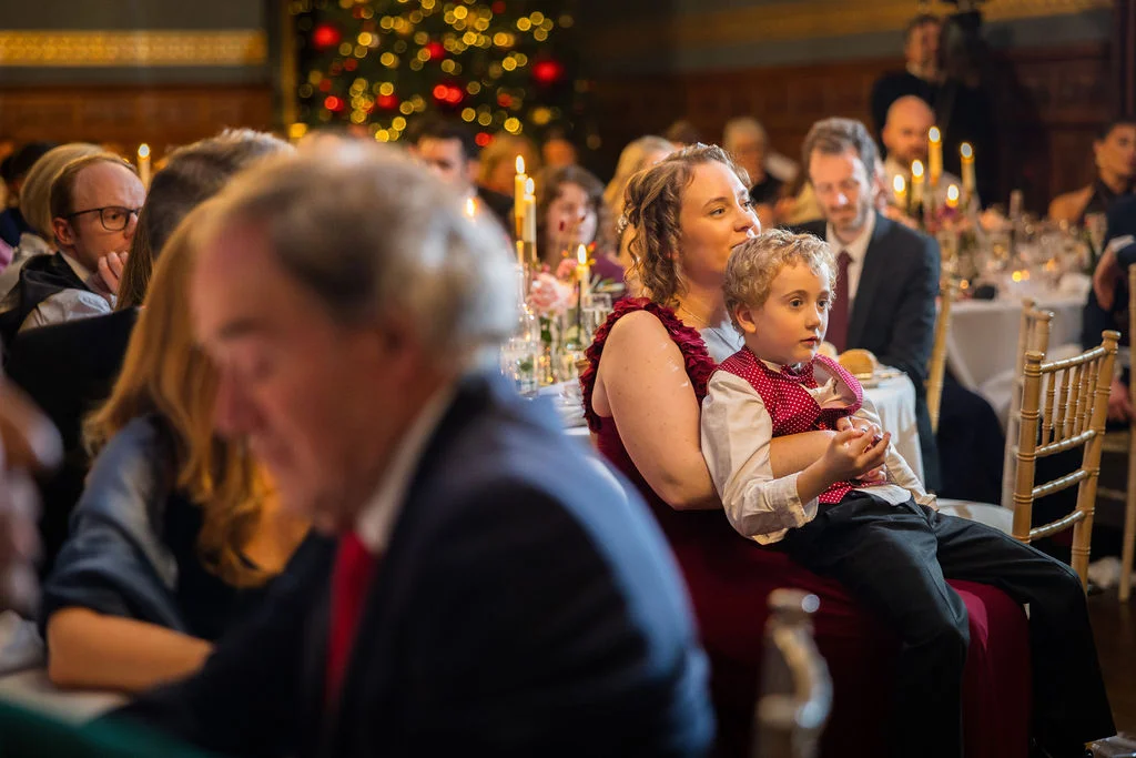 A young boy sits on a guest's lap at a festive wedding reception table decorated with glowing candles and a Christmas tree in the background.
