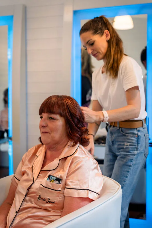 A mature bride with red hair sits in a white chair wearing silk pyjamas while a stylist works on her hair during wedding morning preparations.