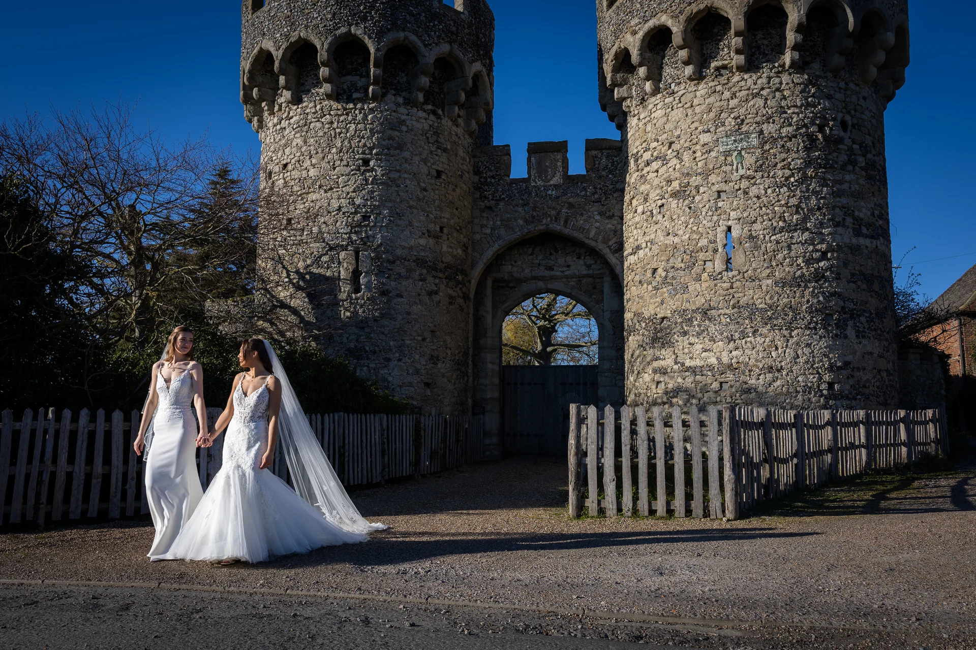 Two brides hold hands and walk together outside a historic stone castle gateway under a clear blue sky.