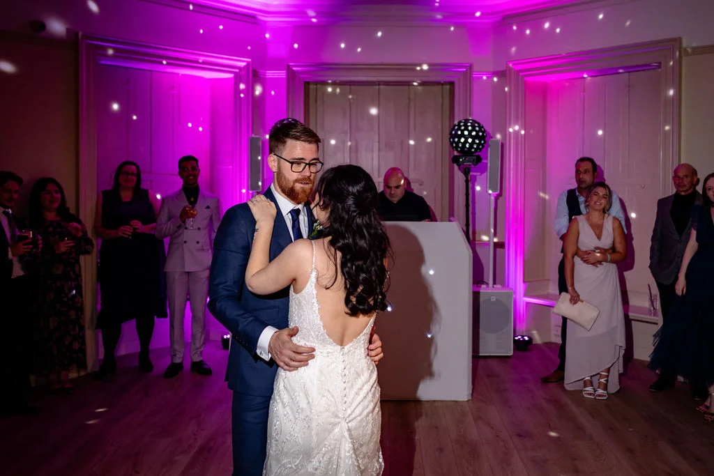 The newlywed couple shares their romantic first dance surrounded by guests in a ballroom illuminated by vibrant pink uplighting and a disco ball.