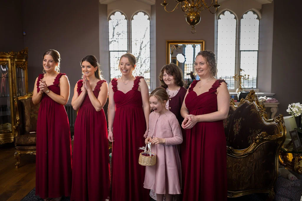 A group of smiling bridesmaids in matching burgundy dresses and a young flower girl stand indoors near arched windows in a stately home.
