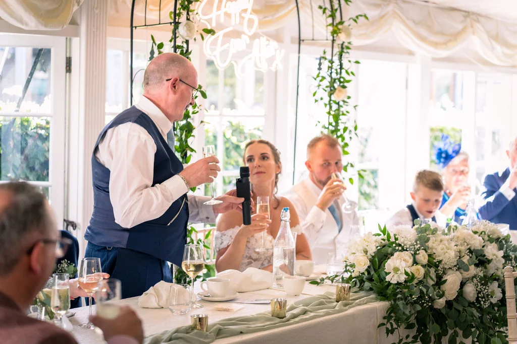 An older gentleman wearing a waistcoat delivers a speech holding a microphone and a glass of champagne at the top table during a wedding breakfast in a light-filled conservatory.