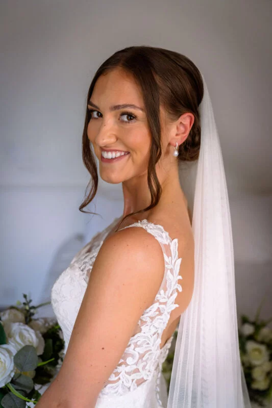 A stunning bride with a low bun and face-framing tendrils smiles warmly over her shoulder while wearing a lace-applique gown and delicate drop earrings.
