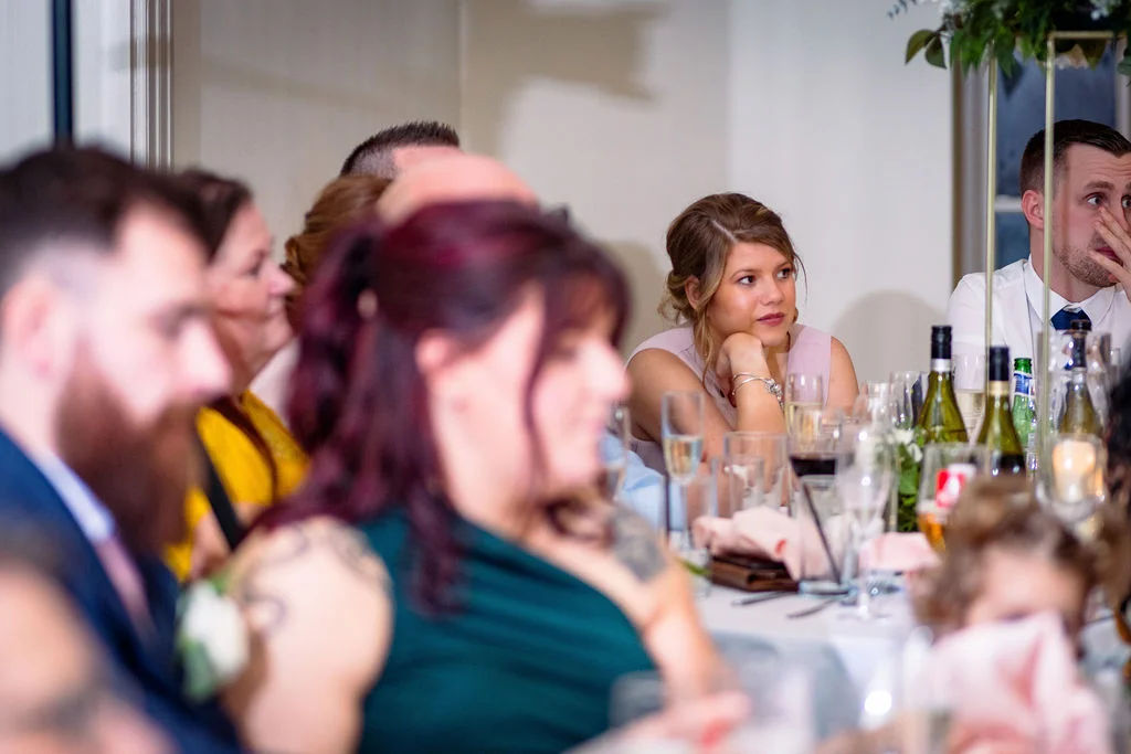 Wedding guests are seated at a reception table, listening intently and reacting during the wedding speeches.