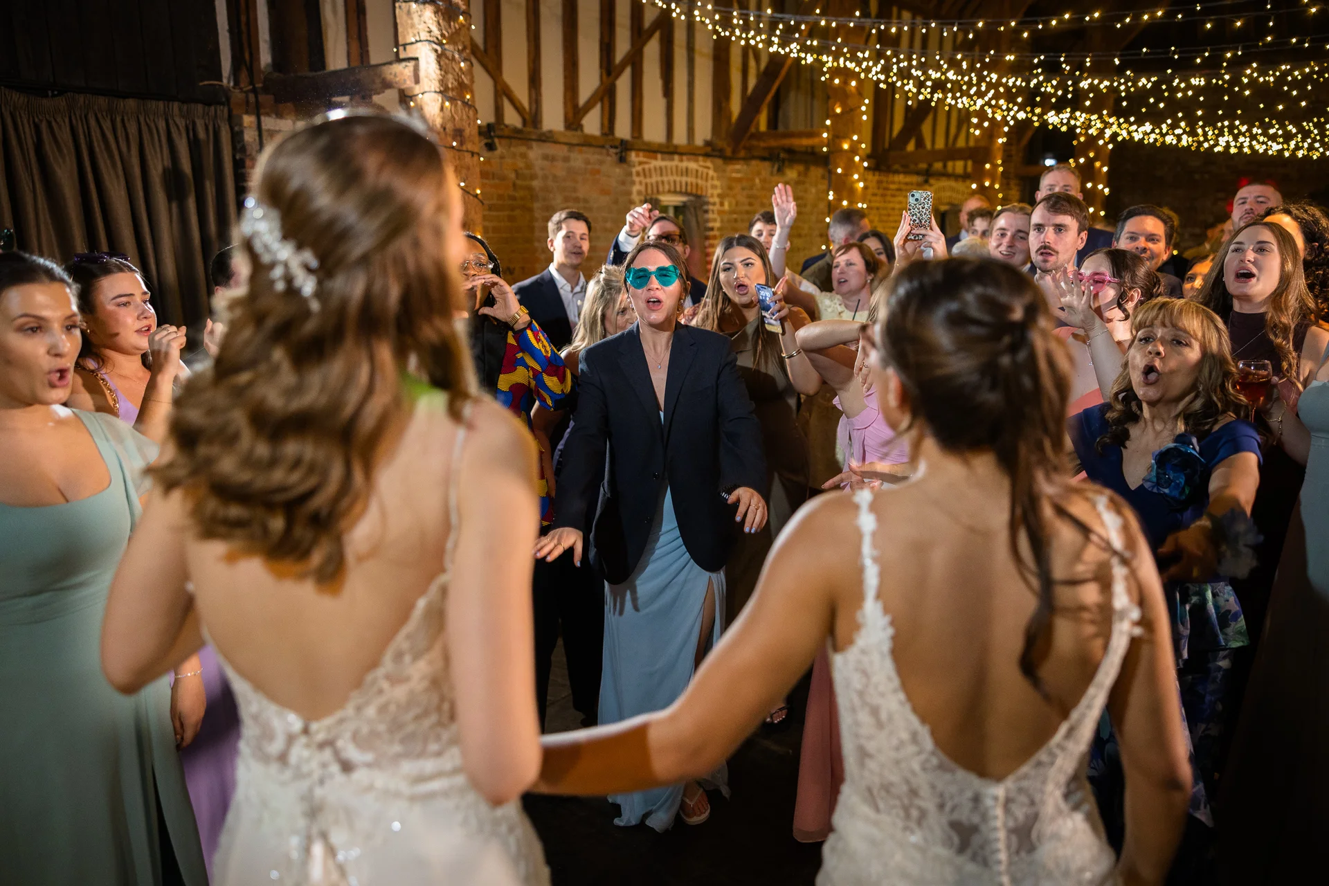 Two brides stand hand in hand facing a cheering crowd of wedding guests inside a rustic barn illuminated by fairy lights.