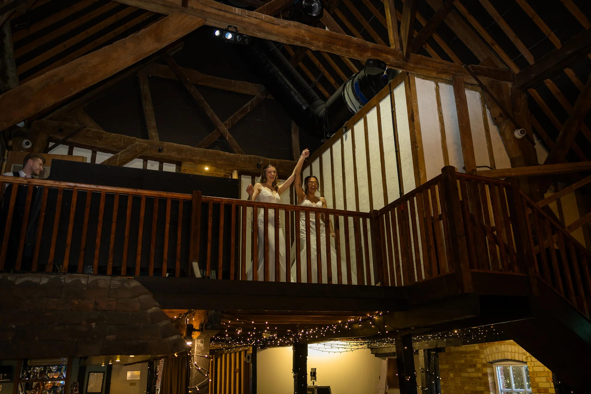 Two smiling brides stand on a timber-framed mezzanine overlooking a rustic barn venue adorned with fairy lights.