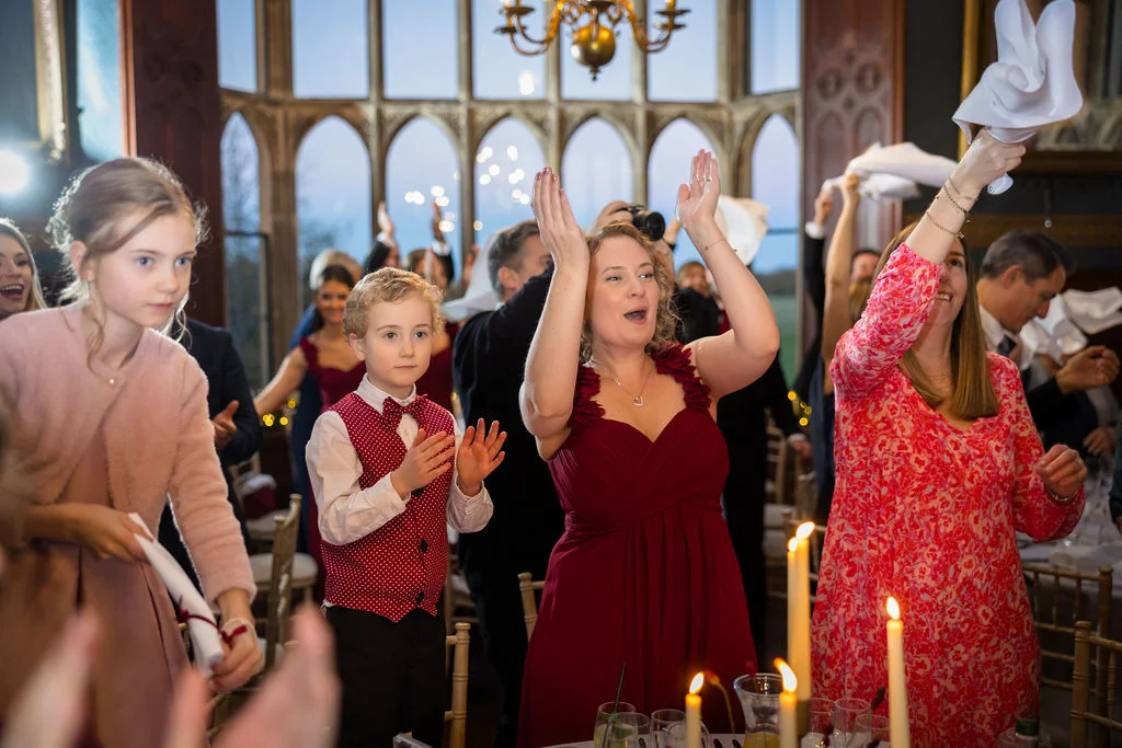 Wedding guests cheering and waving napkins in the air during the reception speeches inside a grand historic room with large arched windows.