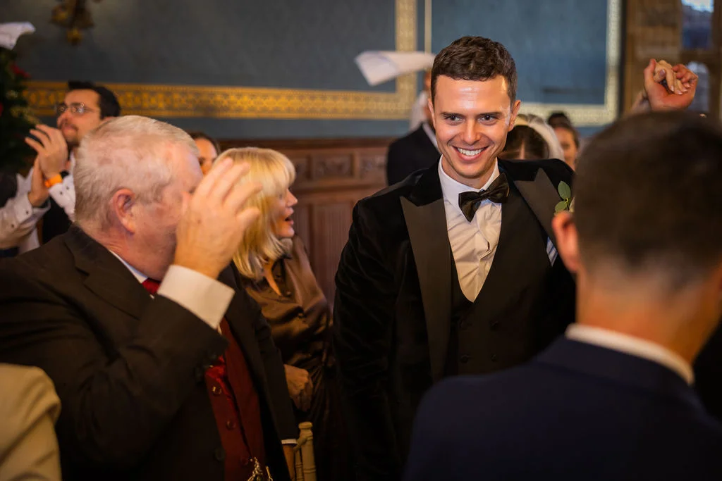 A smiling groom wearing a black velvet tuxedo and bow tie reacts happily as he walks past cheering wedding guests.