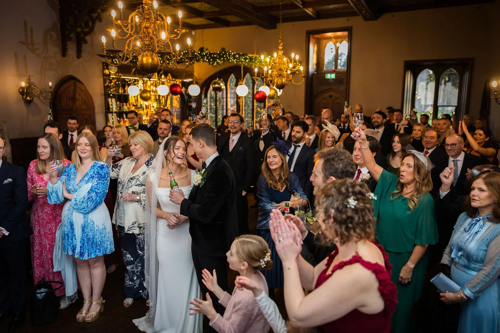 A newlywed couple shares a joyful moment holding drinks while surrounded by cheering guests raising a toast inside a grand historic hall.