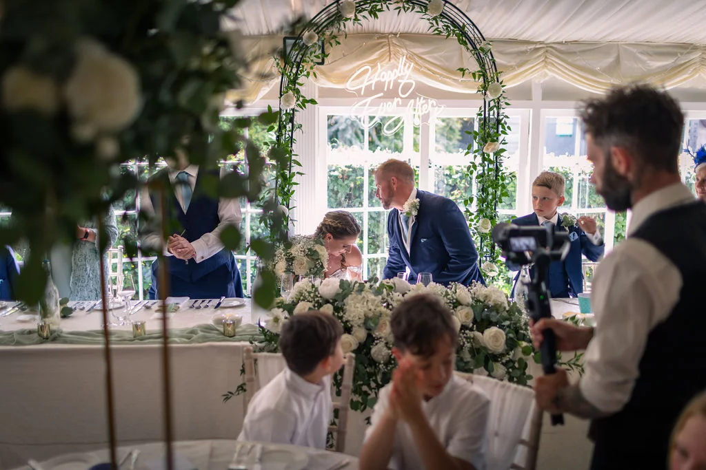 A candid over-the-shoulder shot captures the newlyweds seated at the top table beneath a floral arch and a glowing neon sign while guests and a videographer watch.