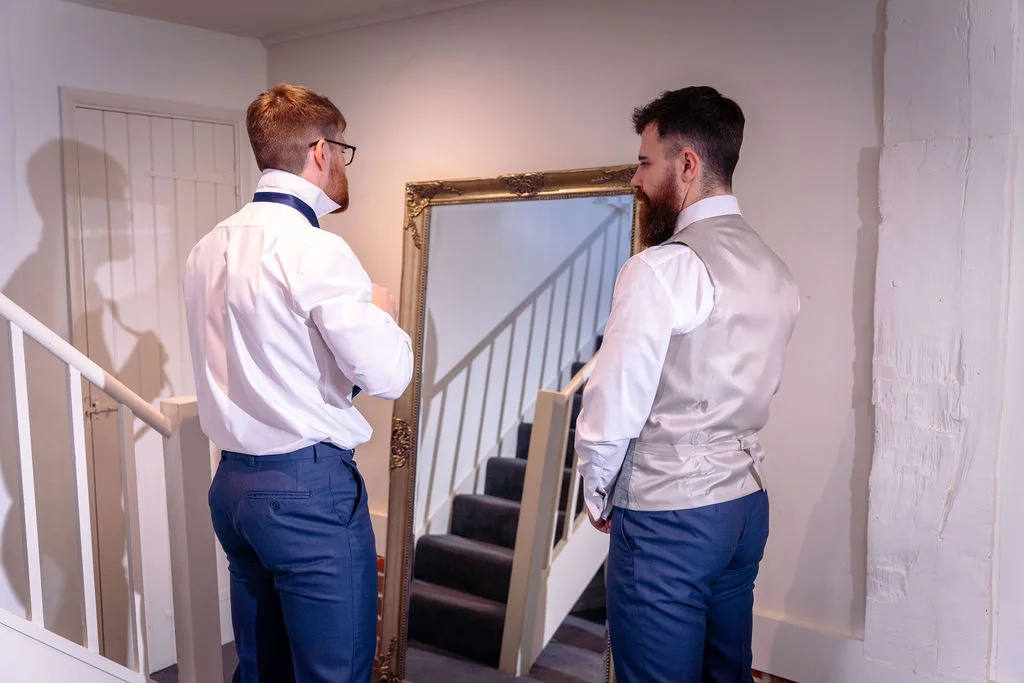 Two grooms stand before a large mirror during morning preparations, adjusting their attire in a bright interior hallway.