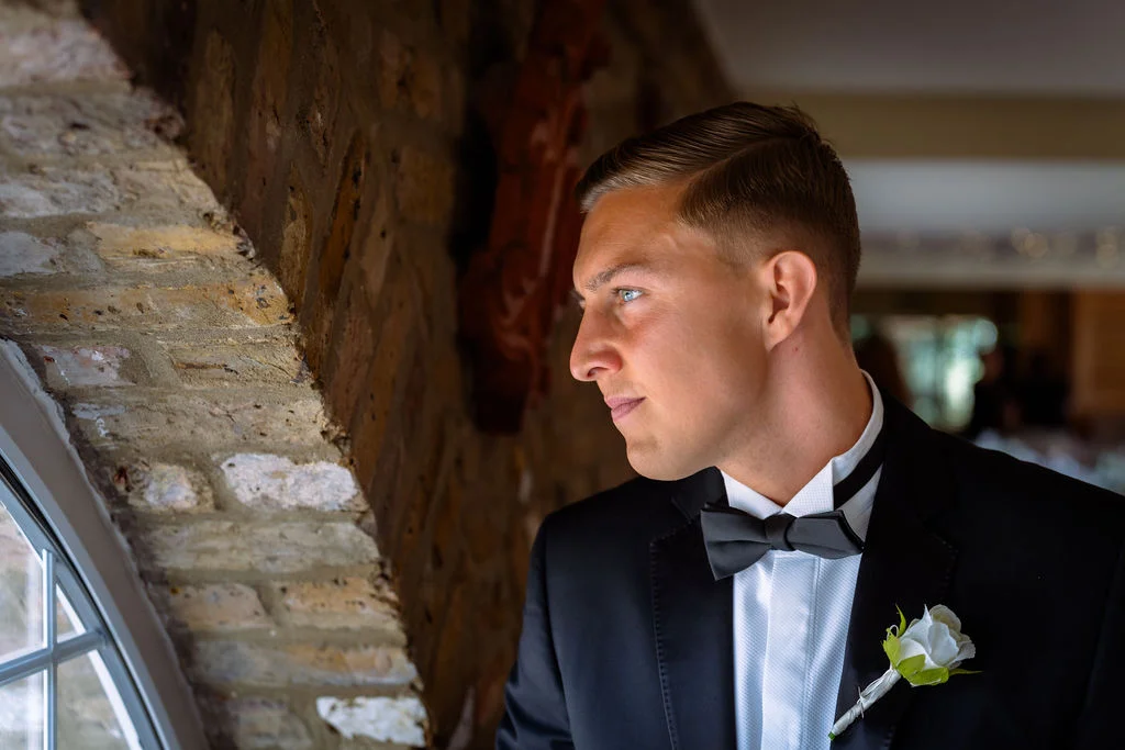A handsome groom in a classic black tuxedo and bow tie stands near a rustic stone archway, looking out thoughtfully with a white rose boutonniere on his lapel.