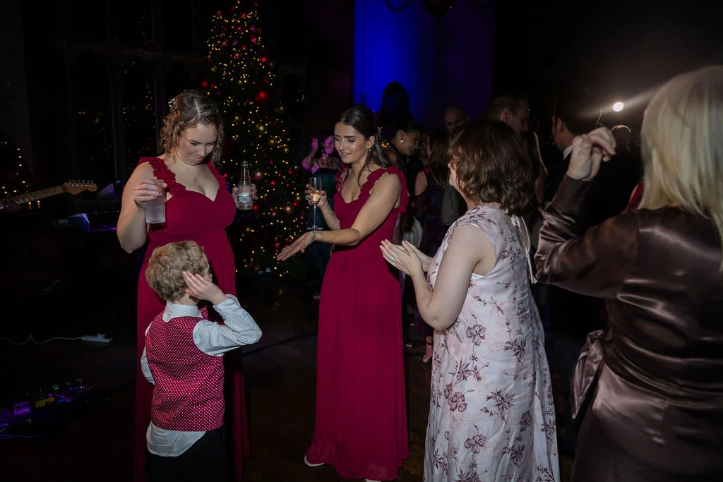 Wedding guests and a young page boy enjoying themselves on the dance floor during an evening reception beside a decorated Christmas tree.