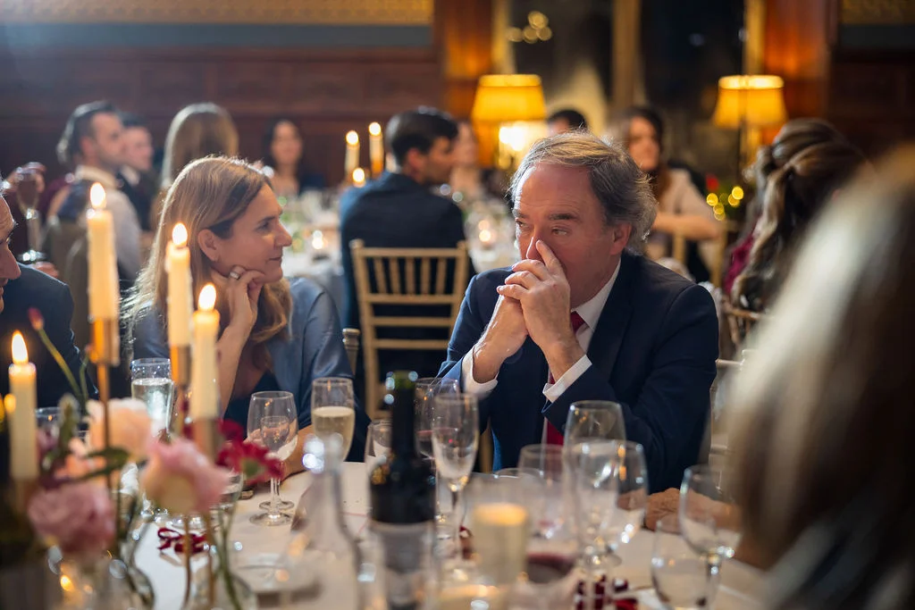 Wedding guests are seated at a candlelit banquet table during speeches, with warm ambient lighting illuminating the wood-panelled dining room.