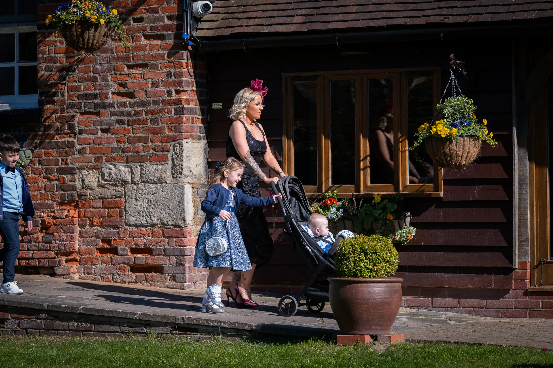 A wedding guest pushes a pram along a paved path next to a rustic brick and wood building on a sunny day, accompanied by a young girl and a boy.