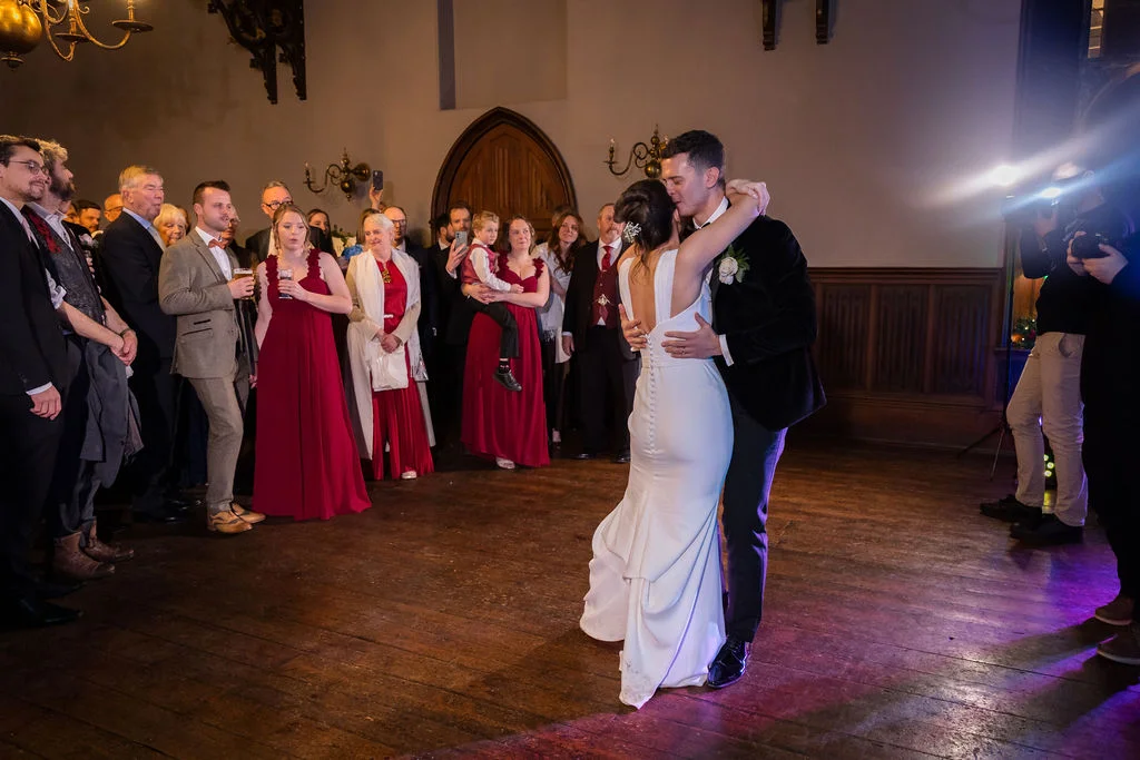 The newlywed couple share their first dance on a wooden floor in a historic hall, surrounded by watching guests.