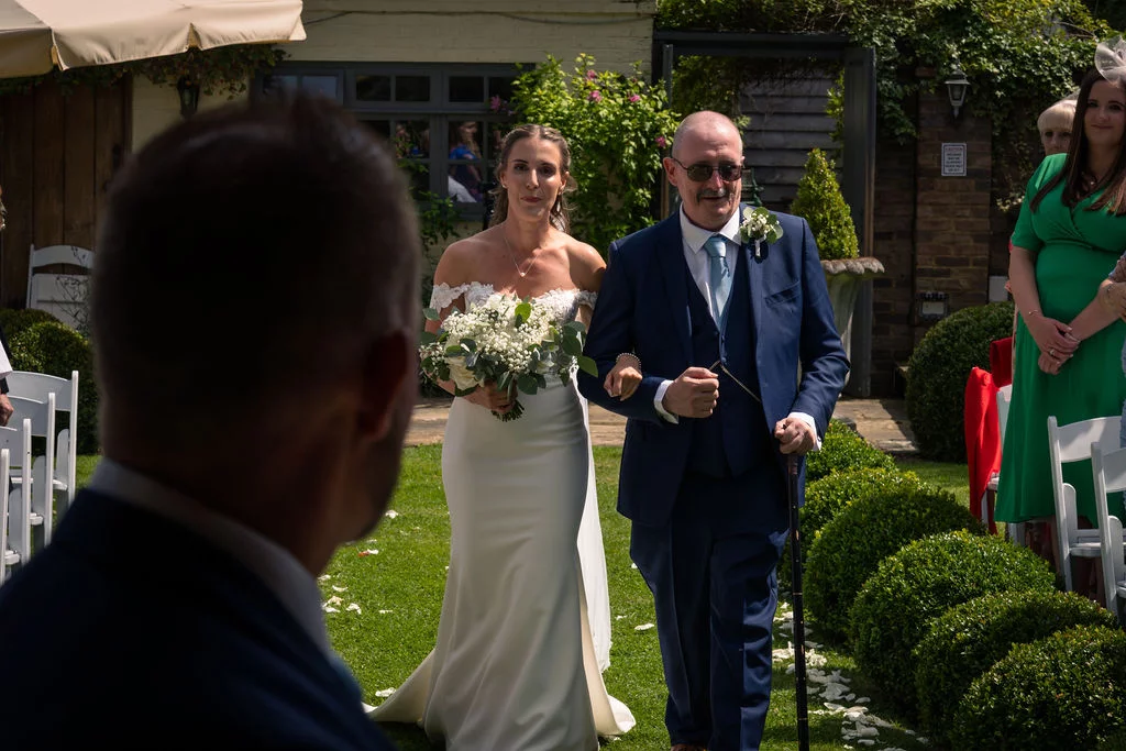 The groom watches from the foreground as the bride walks down the outdoor aisle linked arm in arm with her father.