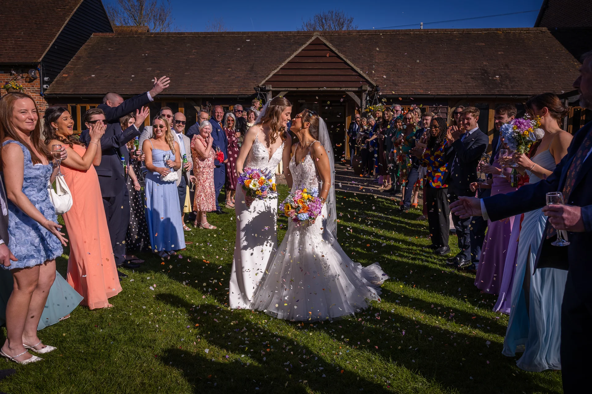 Two smiling brides share a tender kiss on a sunlit lawn as cheering guests shower them with colourful confetti outside a rustic barn.