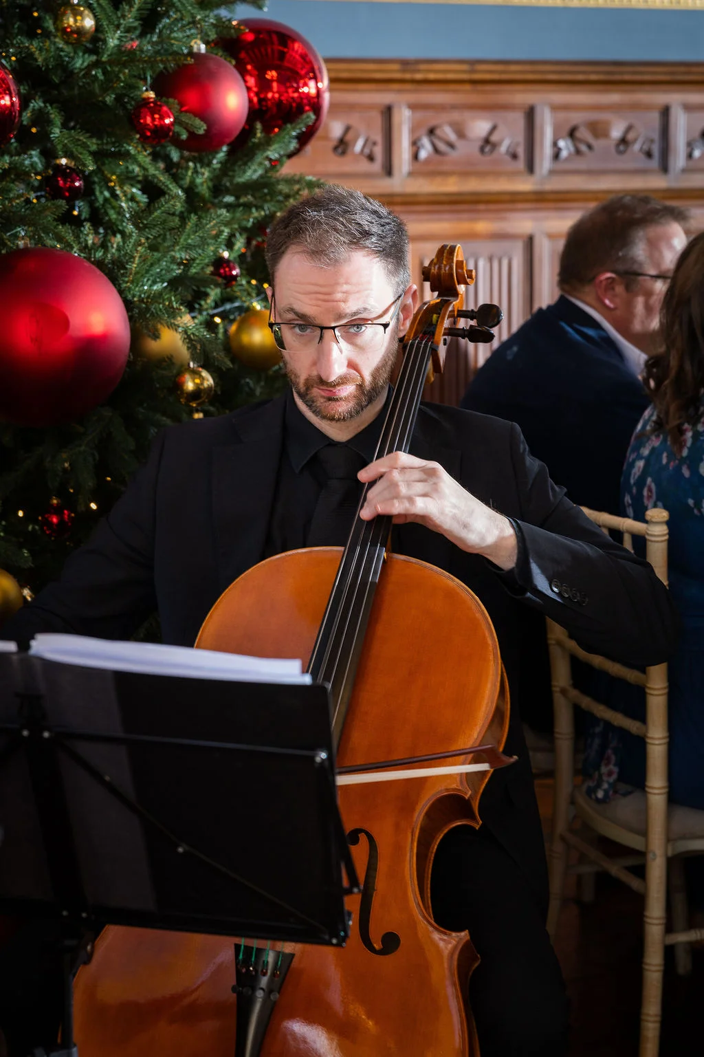 A professional cellist dressed in a black suit performs next to a brightly decorated Christmas tree during a winter wedding reception.
