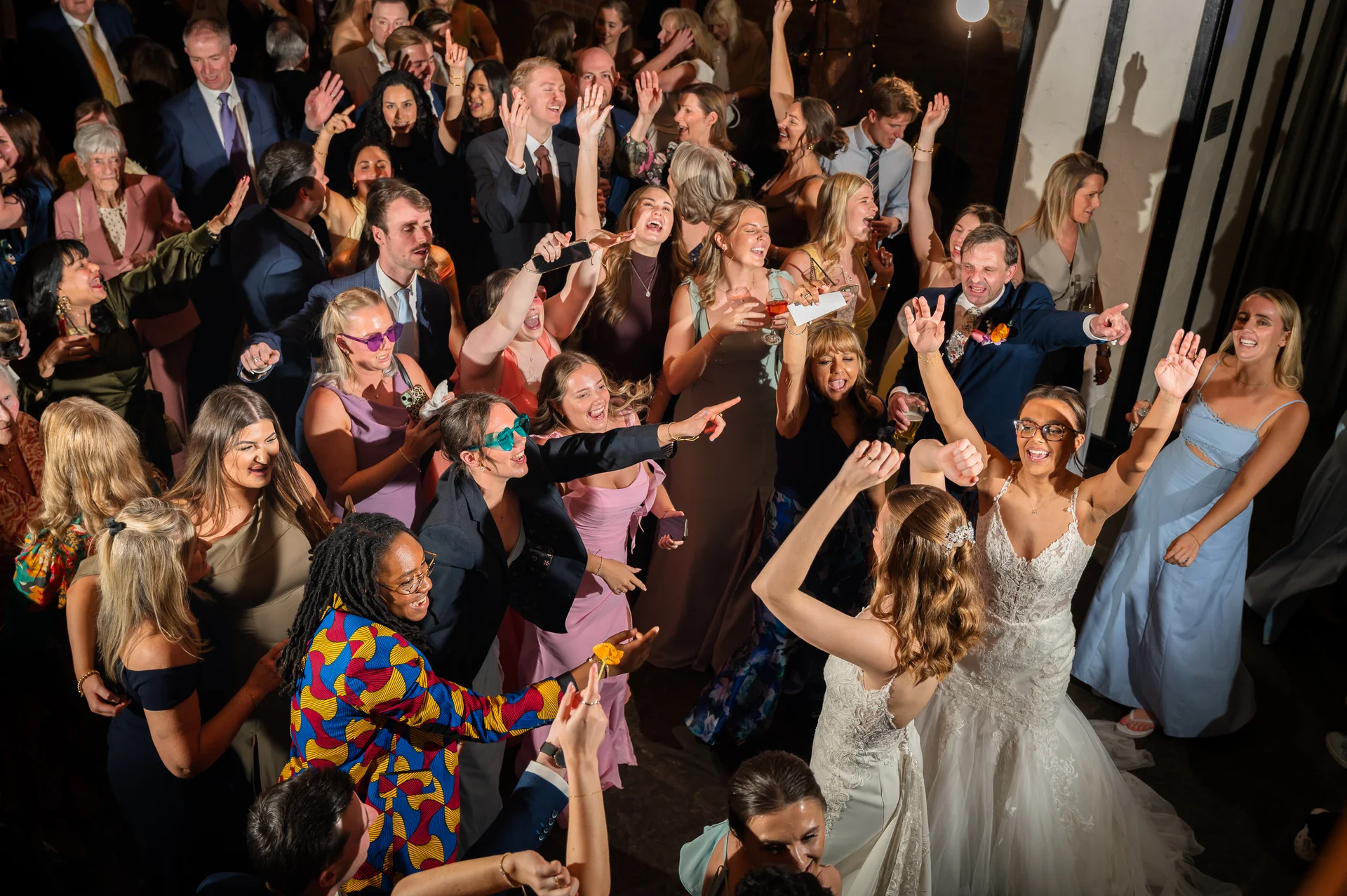 A bride in a white lace gown dances enthusiastically with her guests on a crowded dance floor under warm indoor lighting.