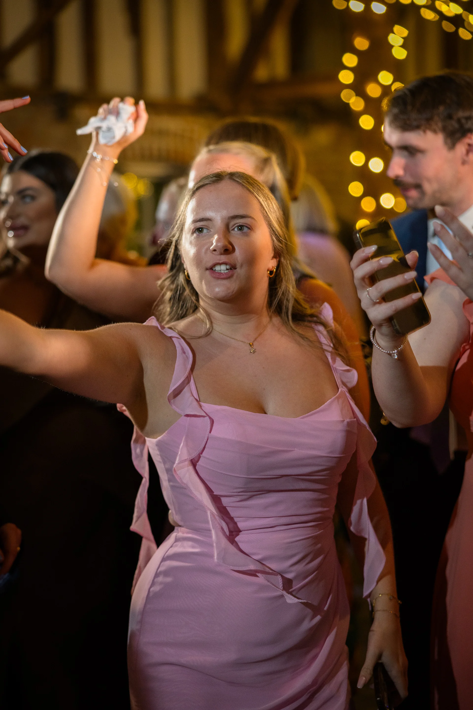 A guest in a pink dress joyfully dances on a lively evening dance floor illuminated by warm fairy lights.