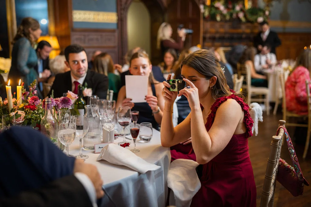 A wedding guest in a burgundy dress holds a disposable camera to take a photo during the wedding breakfast in a wood-panelled dining room.