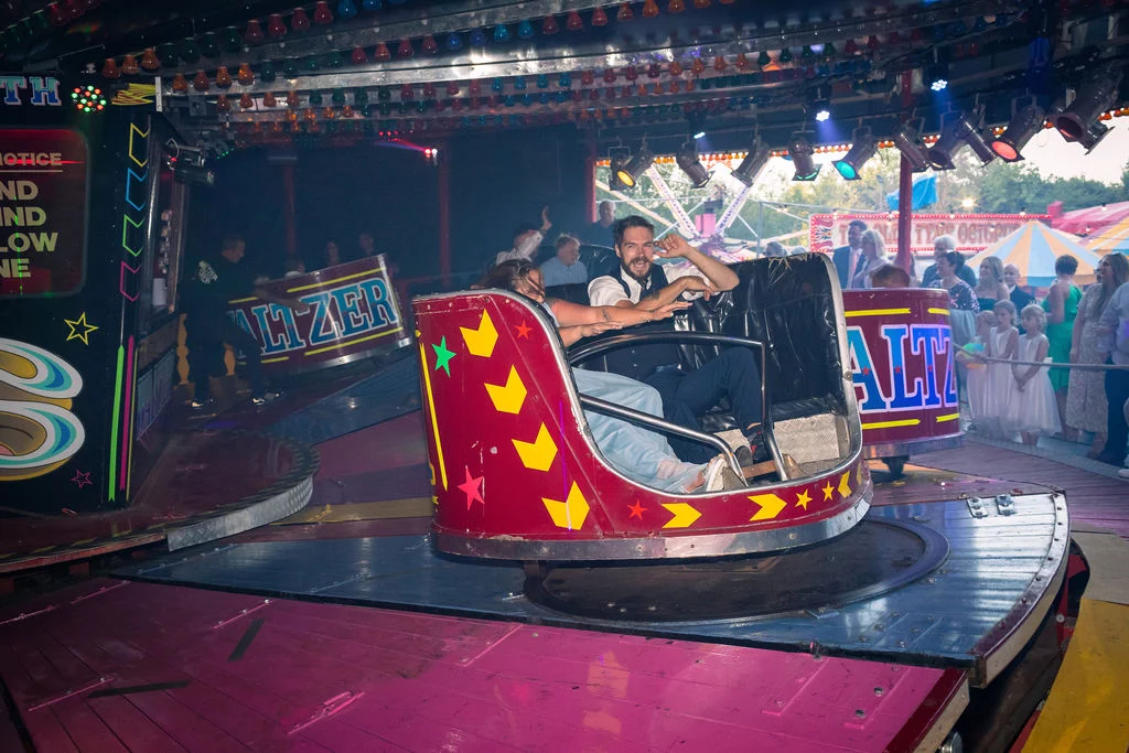 A bride and groom laugh excitedly while riding a brightly lit Waltzer car at a funfair wedding evening reception.