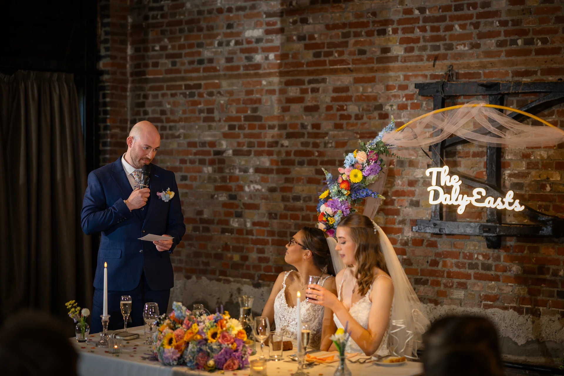 A groom in a blue suit delivers a speech with a microphone at the top table while two brides listen attentively in front of an exposed brick wall decorated with a neon sign and floral arch.