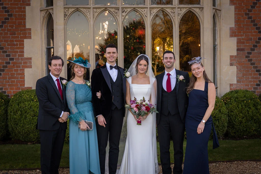 A bridal party and family members pose together outside a historic manor house with large arched windows reflecting a festive Christmas tree.