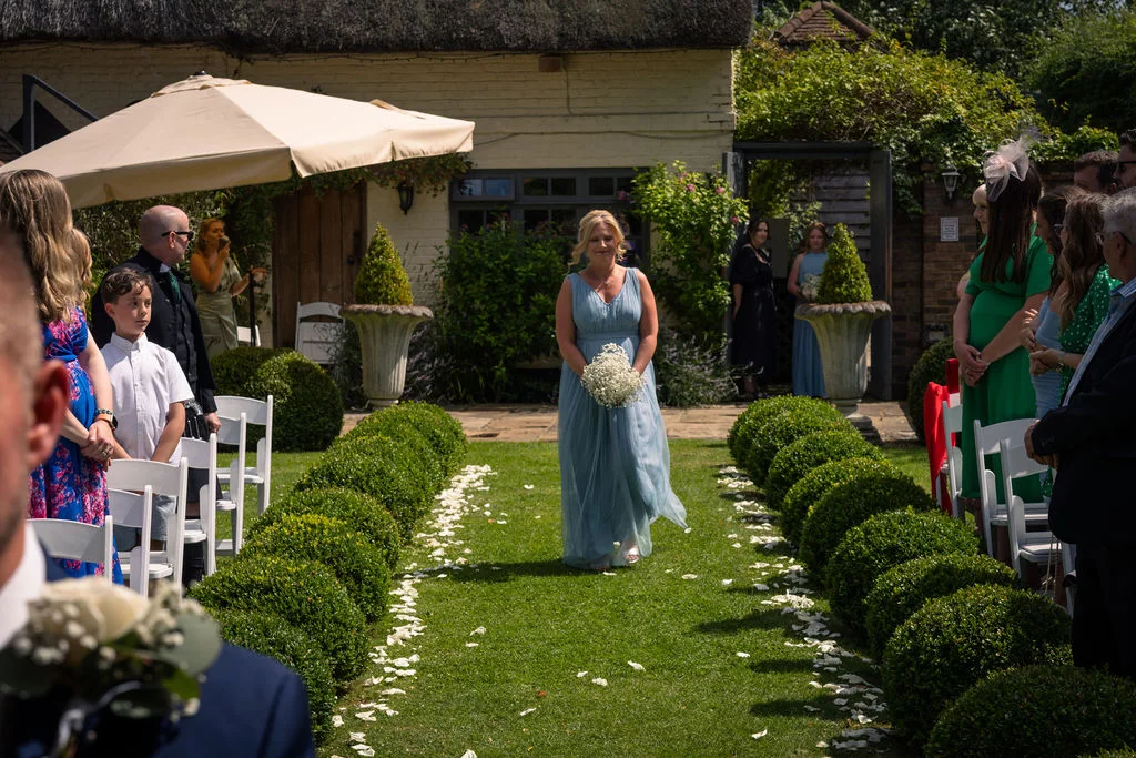 A bridesmaid in a flowing blue gown walks down an outdoor grass aisle lined with topiary bushes and scattered petals during a sunny garden ceremony.
