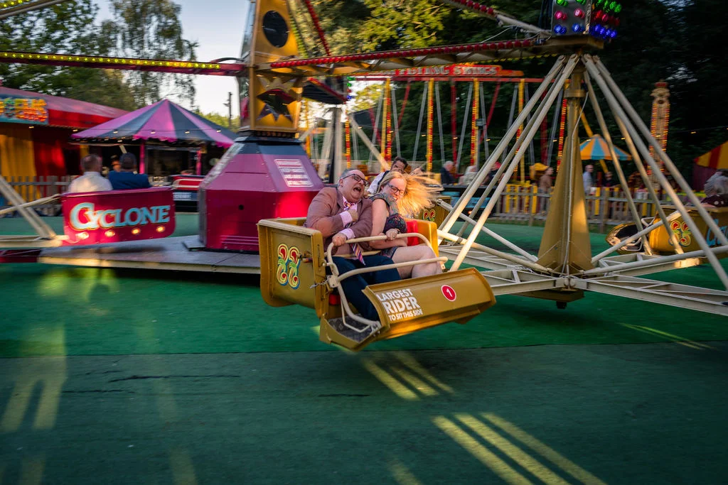 A joyful couple laugh hysterically as they spin wildly in a ride at a vintage funfair wedding reception.
