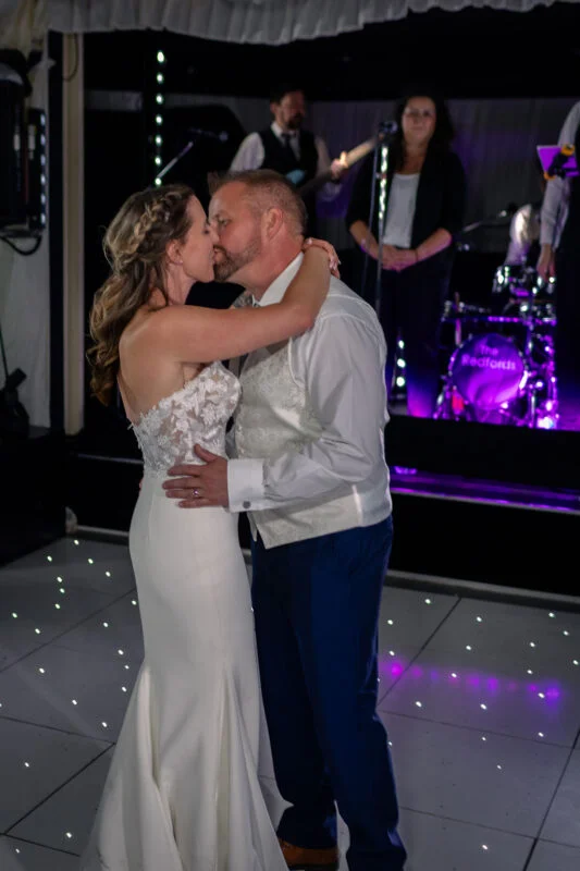 The newlywed couple share a tender kiss on a glowing white dance floor during their first dance, with a live band performing on stage in the background.