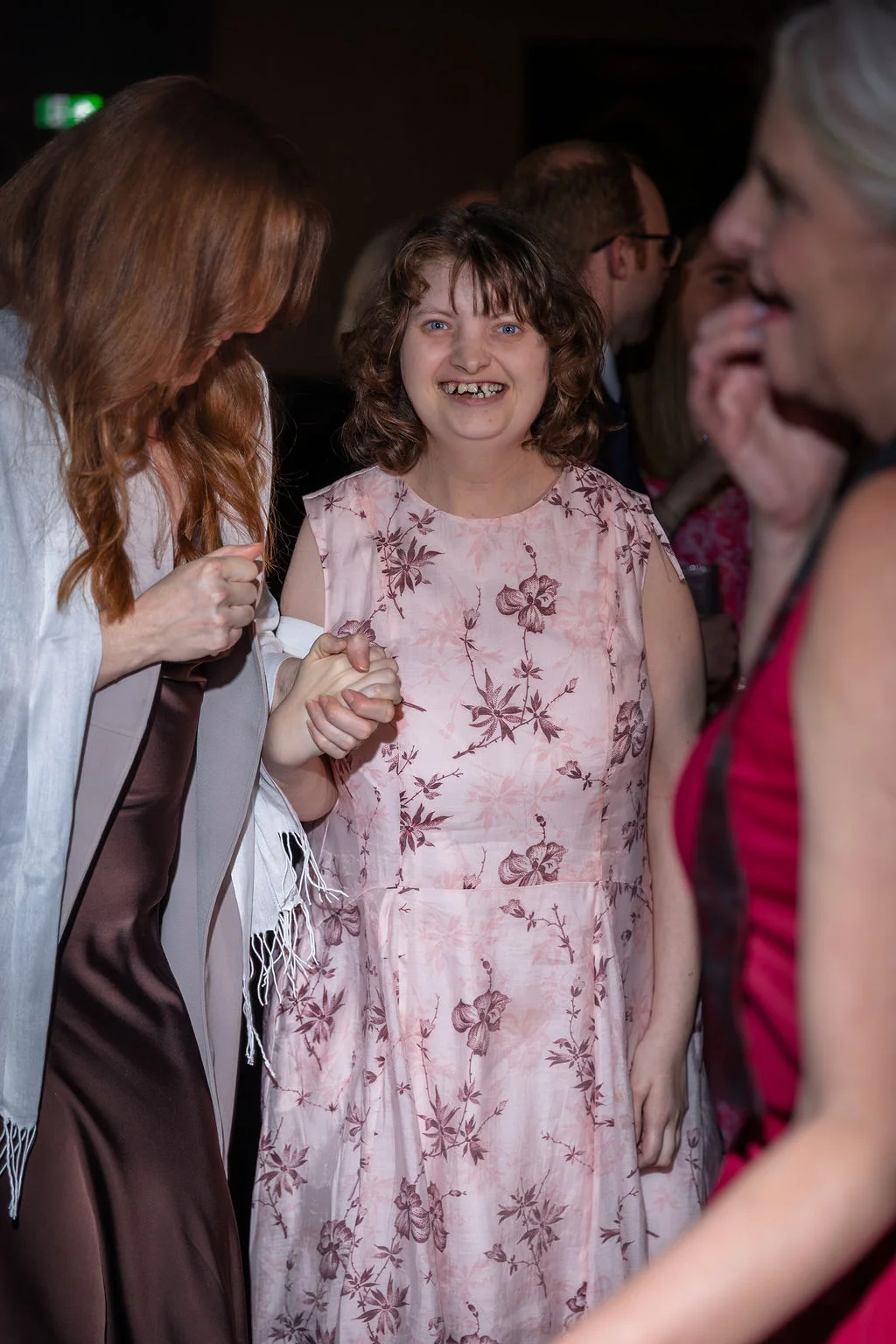 A joyful wedding guest in a pink floral print dress laughs while holding hands with another guest during the evening reception.