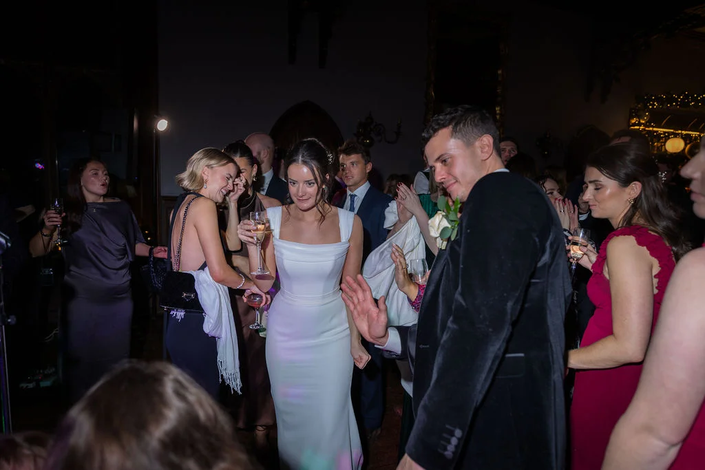 The bride in a sleek white gown and the groom in a dark velvet jacket enjoy the evening reception surrounded by dancing guests.