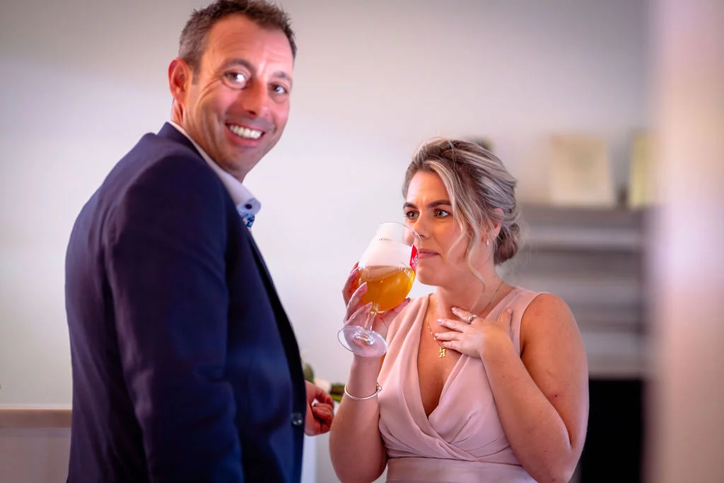 A smiling man in a navy suit looks back towards a woman in a pink dress who is holding a glass of beer during a drinks reception.