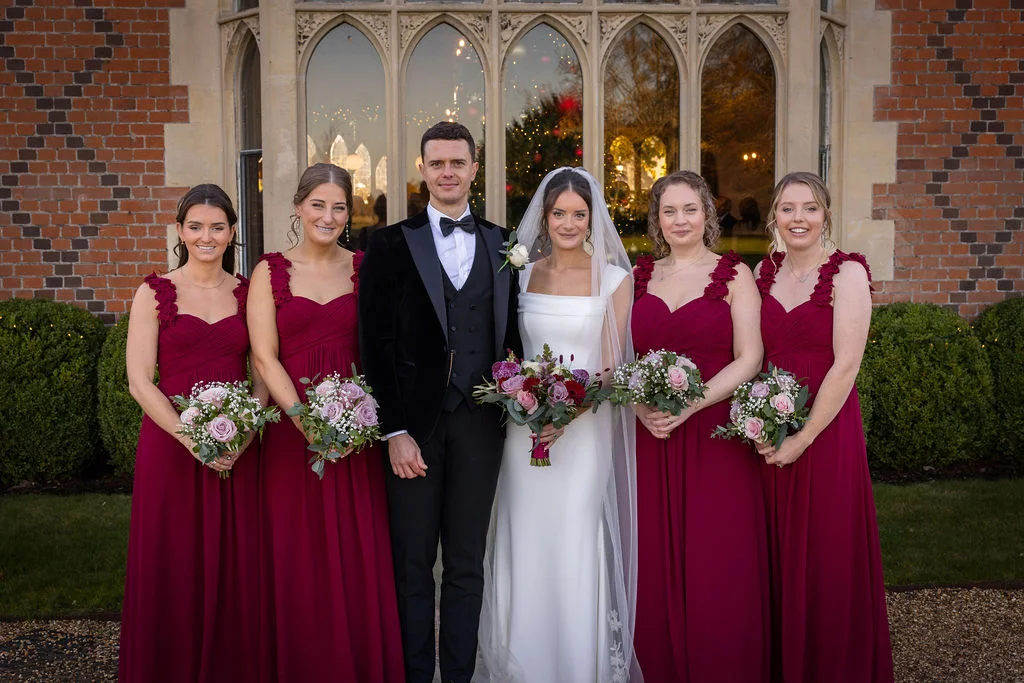 A newlywed couple and five bridesmaids in burgundy dresses pose together outside a historic manor house with arched windows and red brickwork.