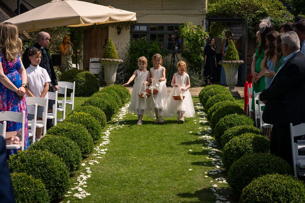 Three young flower girls walk down a grass garden aisle lined with topiary bushes and scattered white petals during an outdoor wedding ceremony.