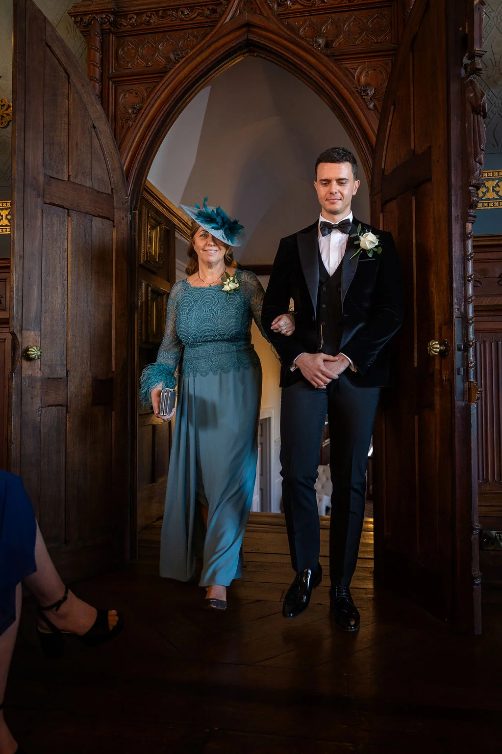 A groom in a black velvet tuxedo walks arm in arm with the mother of the bride through a large wooden doorway in a stately home.