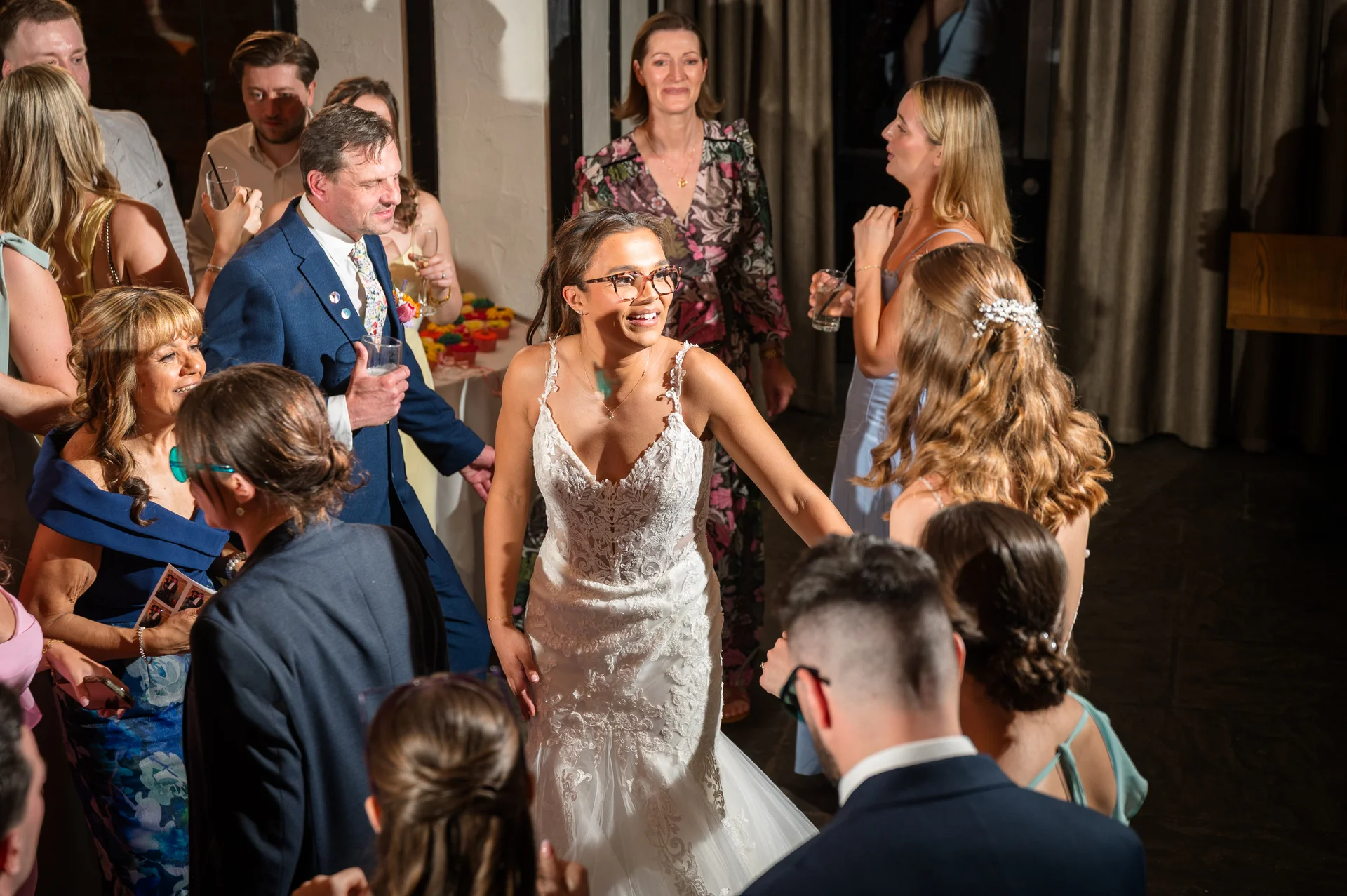 A joyful bride in a lace gown and glasses smiles brightly while mingling with guests during a lively evening wedding reception.