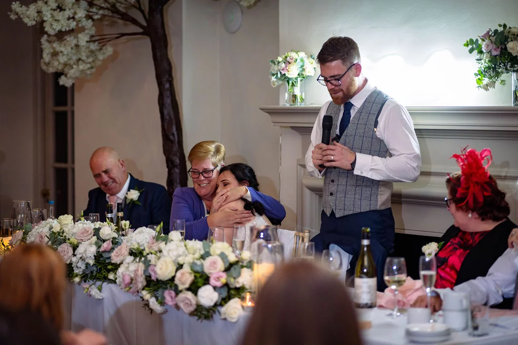 A groom delivers his speech at the top table while a seated guest shares an emotional embrace during the wedding breakfast.