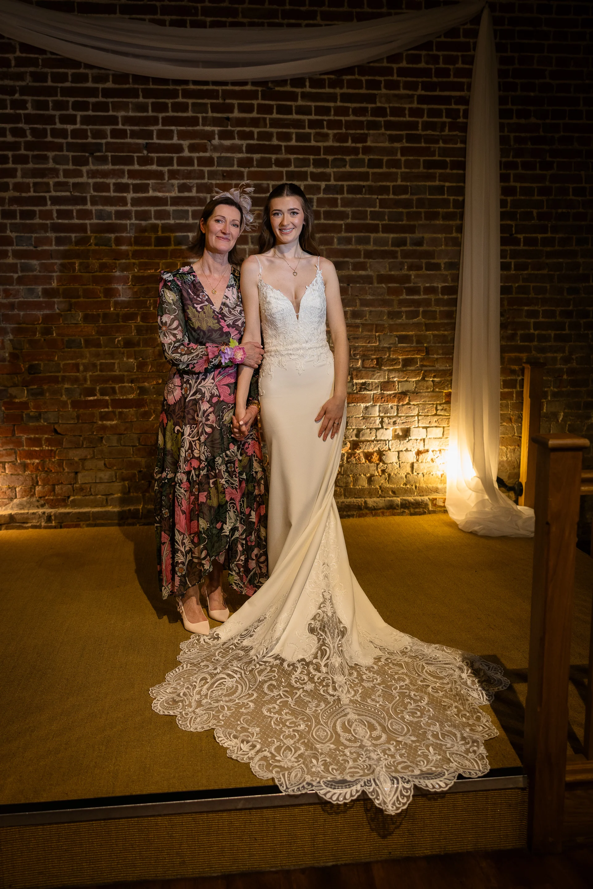 A stunning bride in a sleek V-neck crepe gown with an elaborate lace train poses smiling beside her mother in a floral print dress against an exposed brick wall backdrop.