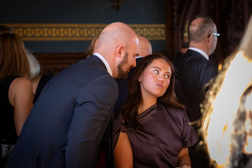A bald guest in a dark suit leans in to whisper something to a woman with long dark hair, who looks away with a playful, quizzical expression.