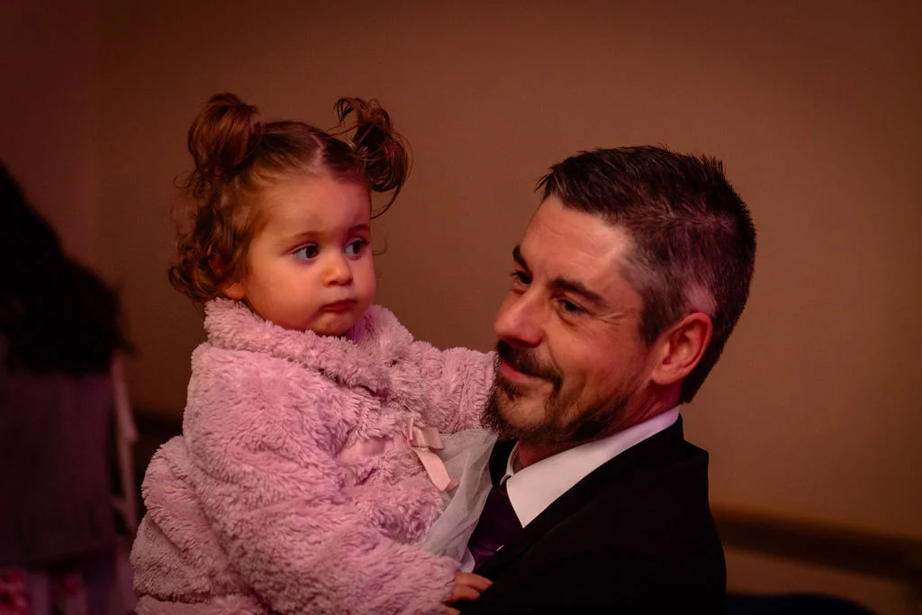 A guest in a dark suit holding a toddler wearing a pink fuzzy coat during the wedding celebration.